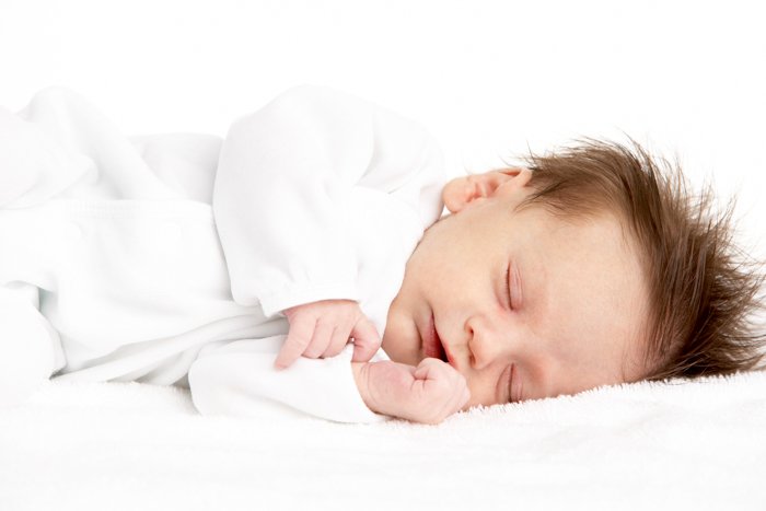 A close-up of a newborn sleeping on his side in a white outfit against a white fuzzy blanket and background