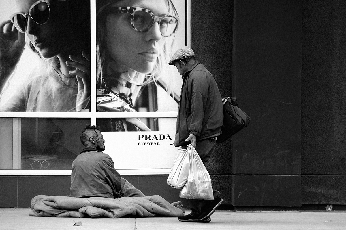 A photo of a person passing a homeless person against a luxury Prada ad