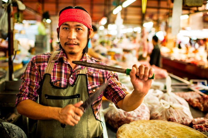 An environmental portrait photo of a market butcher holding knives up in Thailand