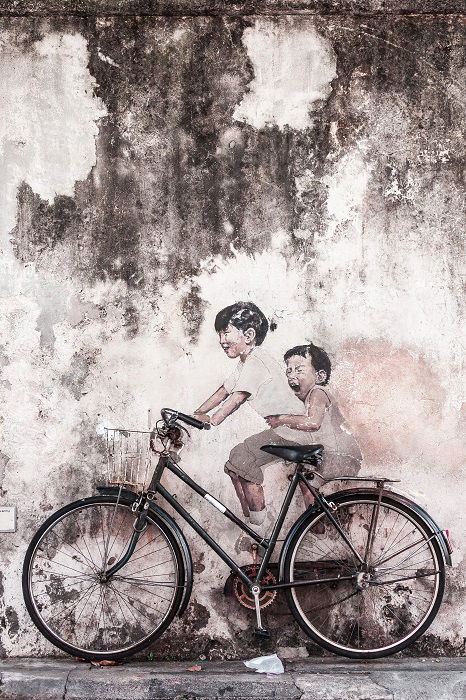 A black-and-white fine-art street photograph of two children sitting on a bike against a wall and laughing