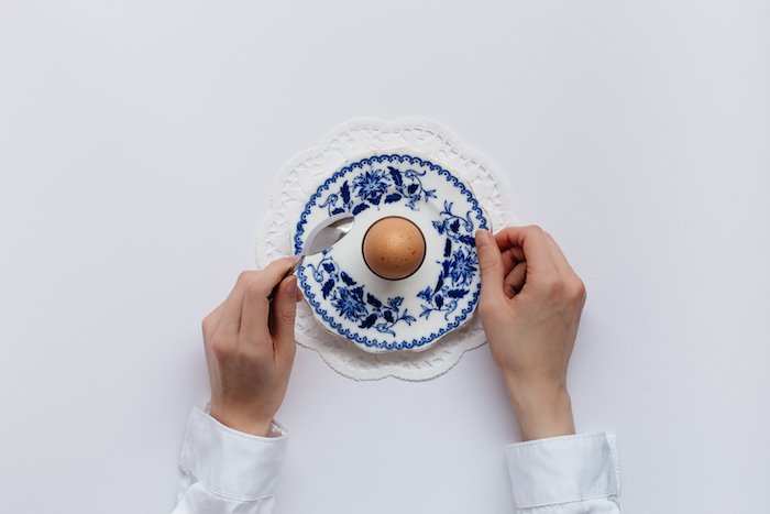 平铺食物摄影:双手拿着餐具，放在一个煮鸡蛋上，盘子上有图案，背景是纯白色