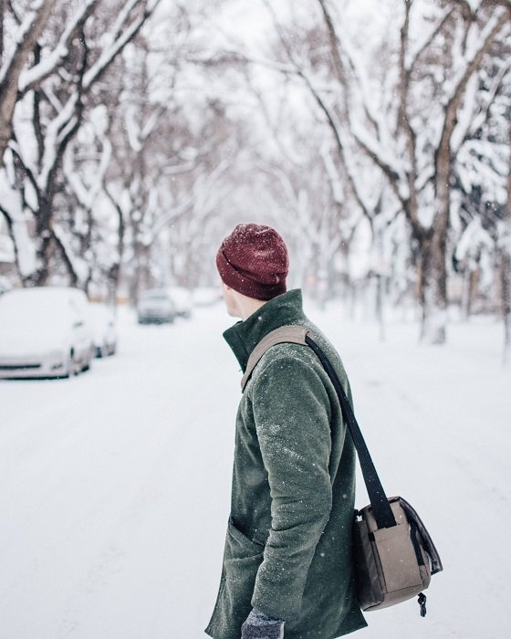 a photographer walks through a snow covered town with a messenger bag over his shoulder