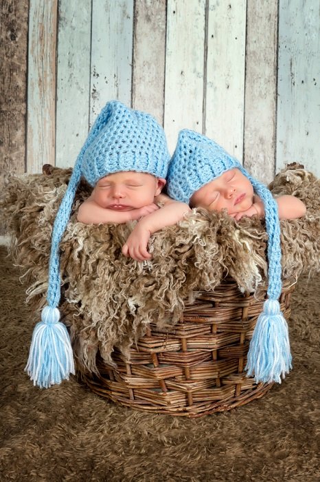 新出生的孪生照片想法：穿蓝色针织的帽子的新出生的双胞胎在一个软的被排行的柳条筐睡觉