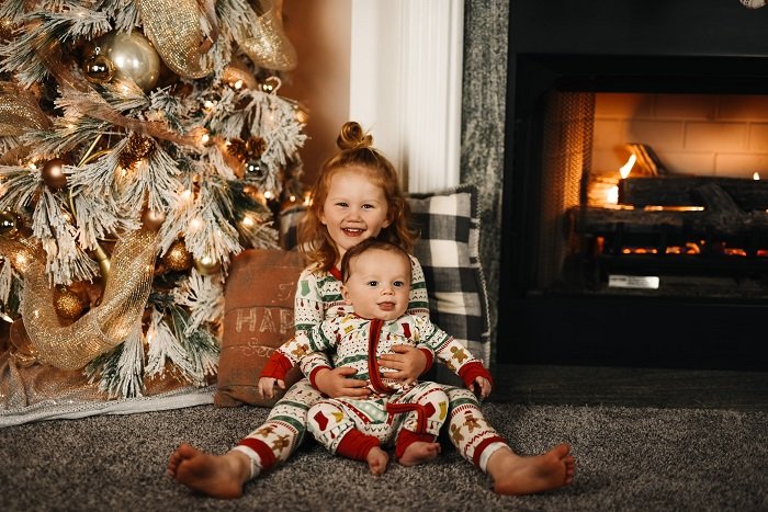 photo ideas for siblings: siblings in matching christmas outfits sitting by a christmas tree