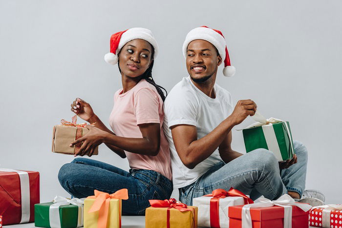 Christmas couple photoshoot ideas: a couple in Santa hats sit back to back and open gifts together