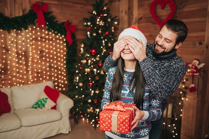 Christmas couple photoshoot tips: a man holds his hands over his girlfriends eyes so she can't see the present she's holding 