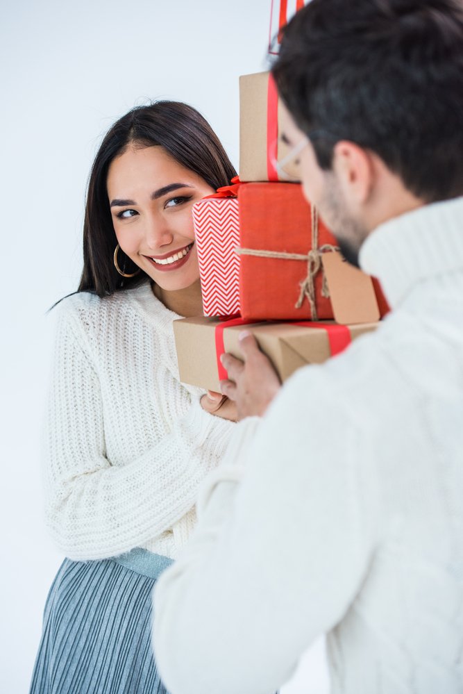 Christmas couple photoshoot tips: a woman looks at her boyfriend from behind wrapped Christmas presents 