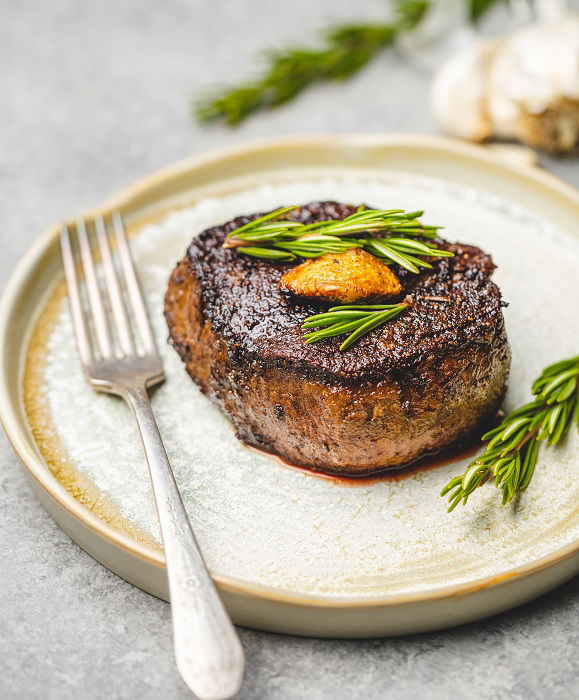 A piece of cooked steak garnished with rosemary on a plate used as a food photography prop