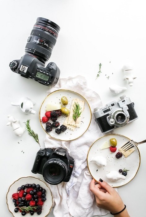 a flat lay photo of cameras and delectable food sitting on a white table