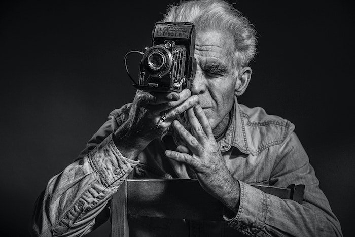 An older man holding up a film camera to his eye taken by Paula Morin (Unsplash.com)
