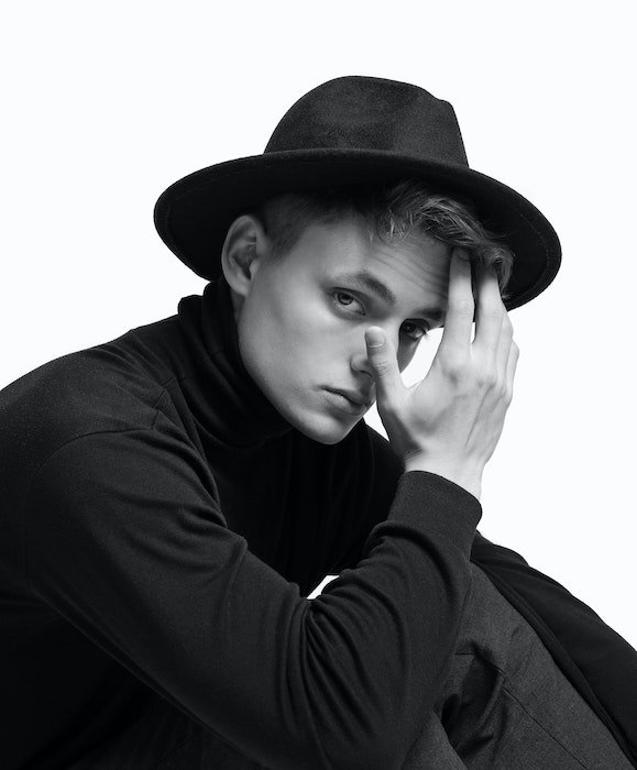 Black and white studio photo of a model wearing a hat and touching their face as an idea for male poses