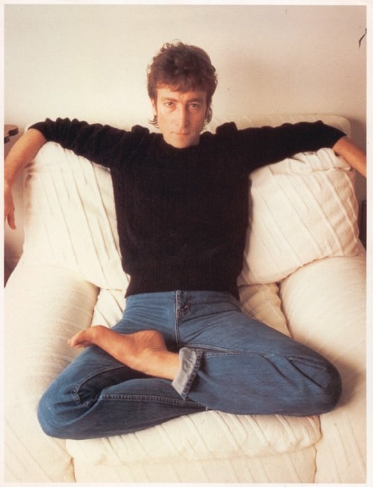 portrait of John Lennon sitting crossed legged on a white couch 