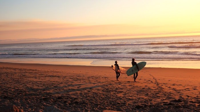 Two surfers walk towards the water at dusk
