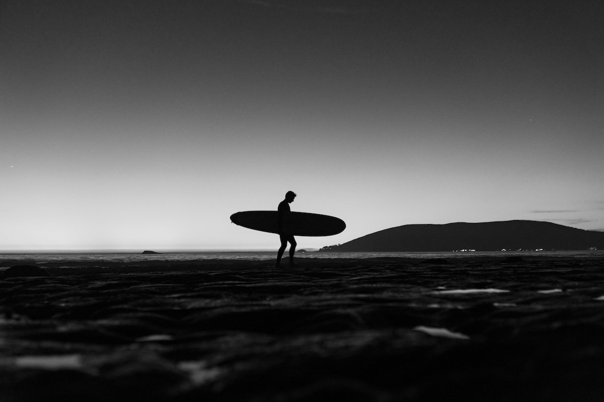 A black-and-white silhouette of a surfer carrying a surfboard from the water as an example of surf photography