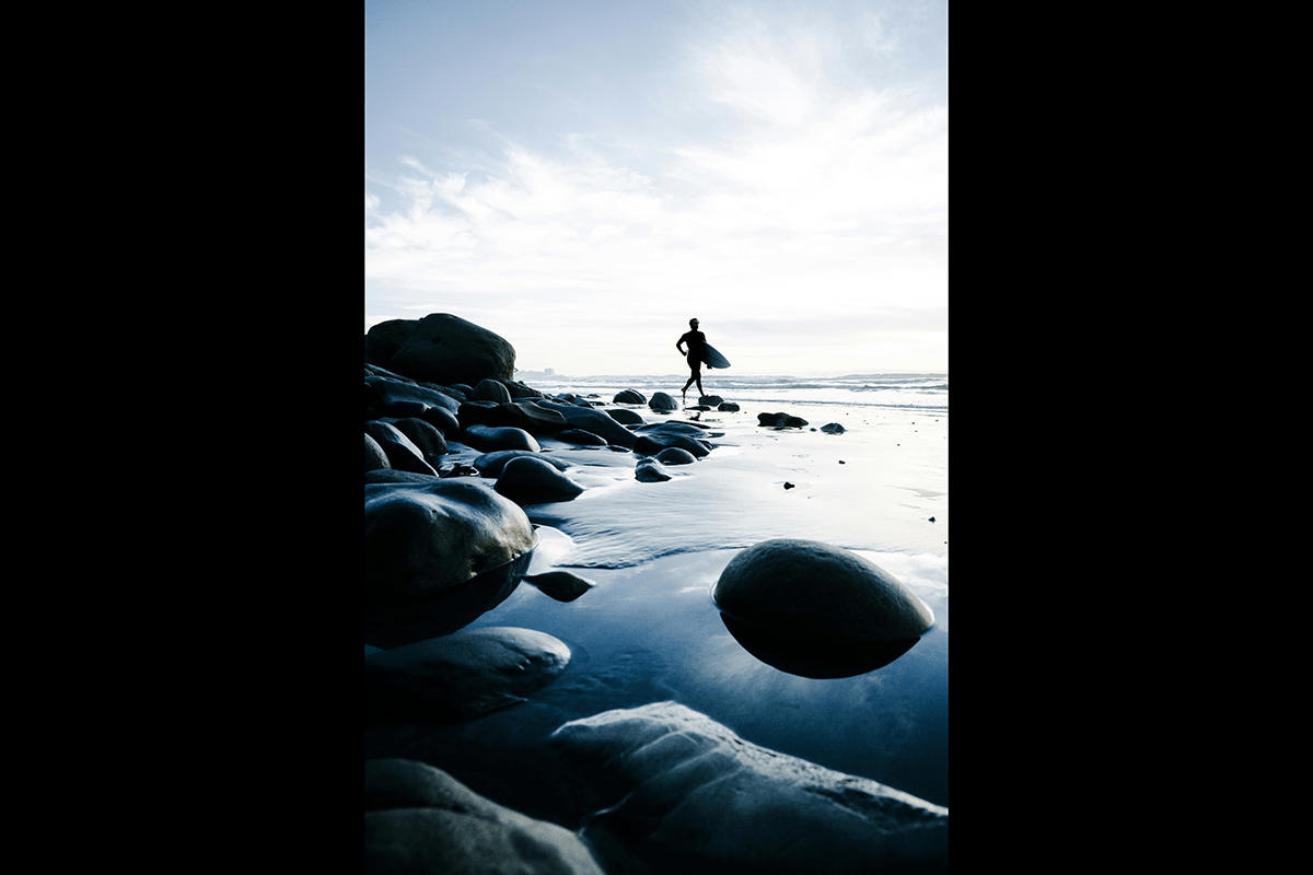 Environmental shot of a surfing running to the ocean with a surfboard with rocks in the foreground as an example of surf photography