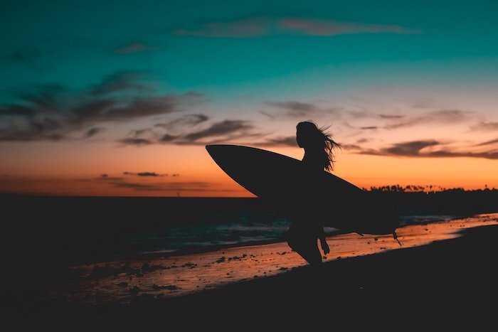 A surfer holding a surfboard is silhouetted against a sunset