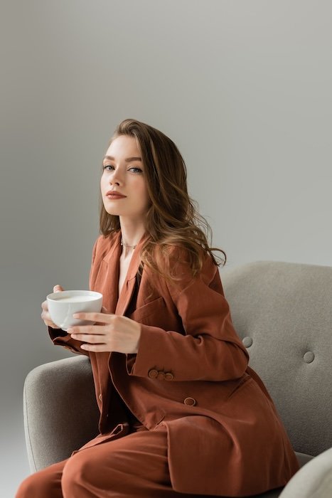 Stock photo of young woman in terracotta suit looking at camera while holding cup and sitting on modern armchair isolated on grey