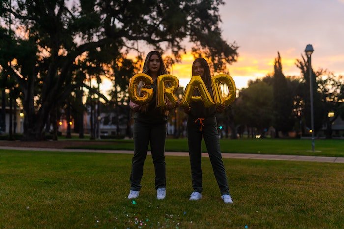 Two girls standing in a park and holding balloons that spell out "grad"