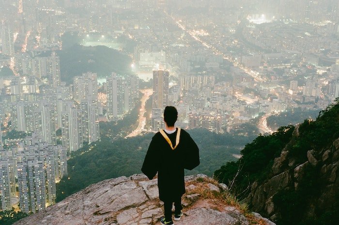 Person in a black graduation gown standing and looking down on a city from a cliff