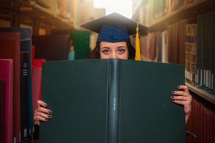 Senior high-school peeking over a large book with a graduate cap on for a senior photoshoot