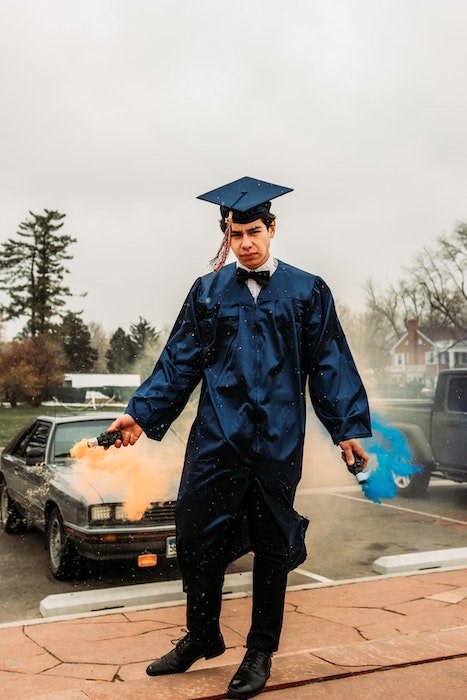 Unique senior picture idea of a senior high-school graduate in cap and gown holding colored smoke bombs his hands