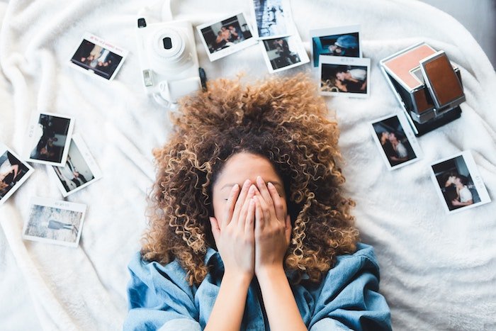 Woman lying on a bed with her hands over her face and Polaroid prints around her