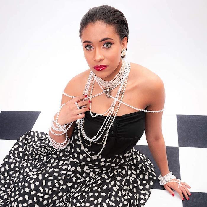 Model in a dress and jewelry sitting on a checkered floor against a white background