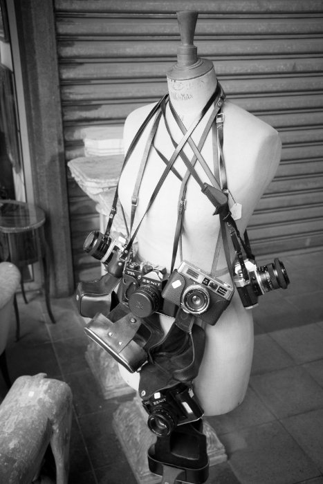 A black and white photo of a mannikin with many vintage cameras around its neck 