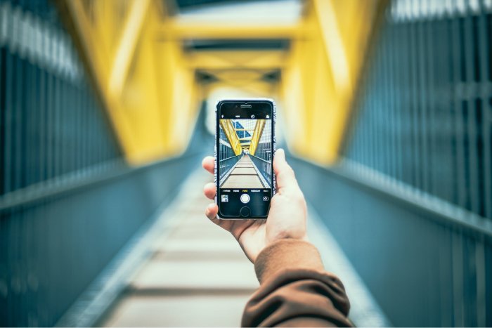 A hand holding up a phone against a blurry background to take a picture of an urban walkway