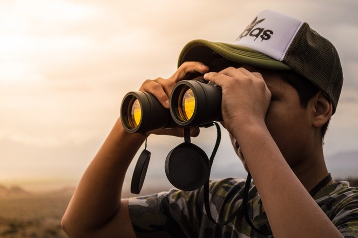A person with a hat and a camouflage shirt on looking through a pair of binoculars for wildlife