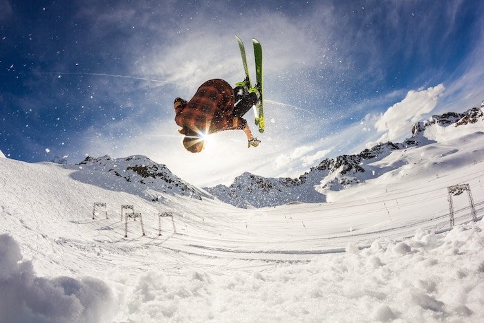 A skier doing a back flip over snow under a blue sky by mountains taken with a slow-motion camera