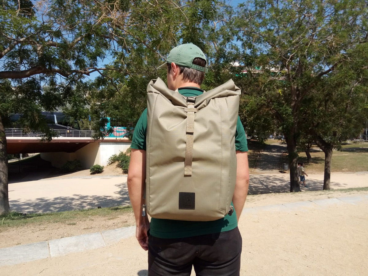 A person wearing a cap and jacket, standing outdoors, with their back to the camera, showing a large tan backpack. 