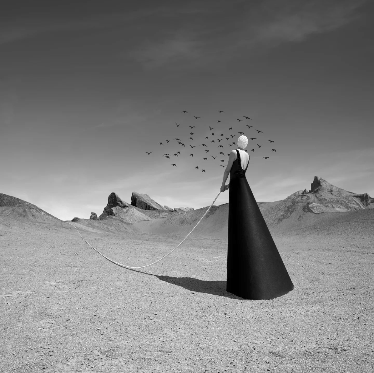 Black-and-white image of a woman in a dress and cap, holding rope in a mountainous desert landscape with birds flying above