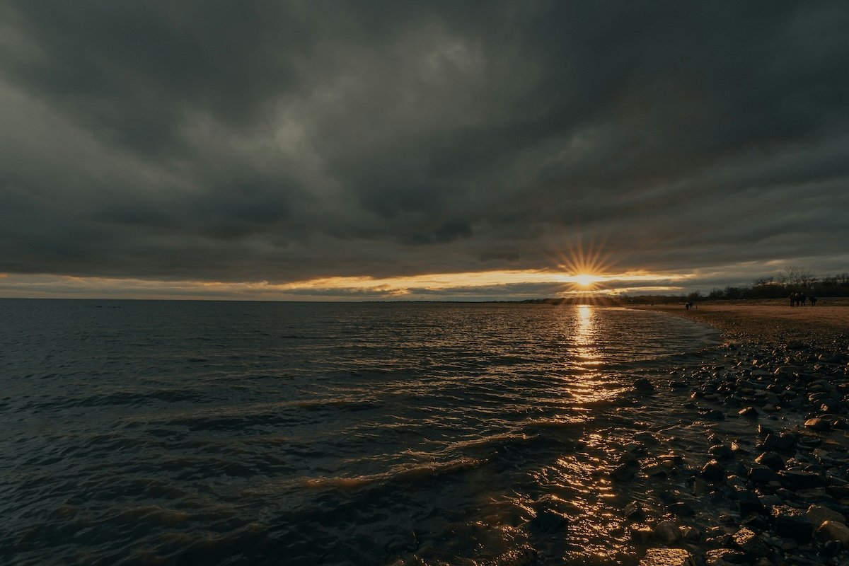 A heavily clouded beach and water seascape with a sunburst sunset on the horizon taken with a narrow aperture