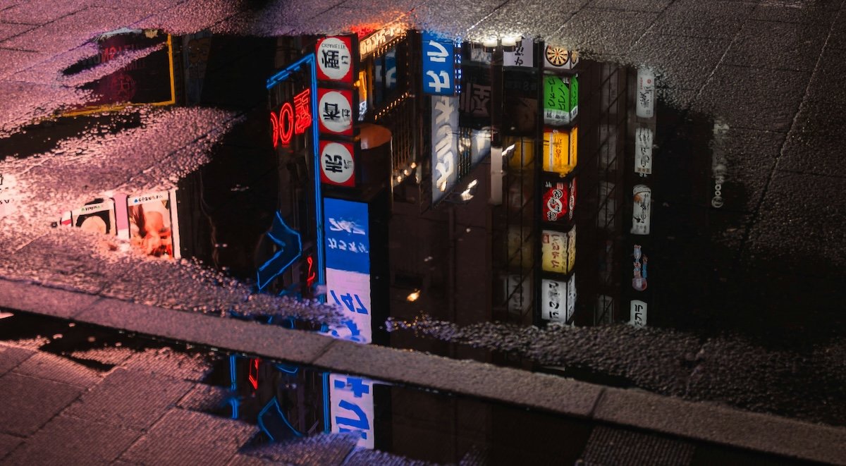 A reflection of neon signs in a rain puddle on pavement at night shot with a wide aperture