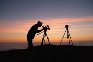 Man setting up two tripods on a mountain top at dusk