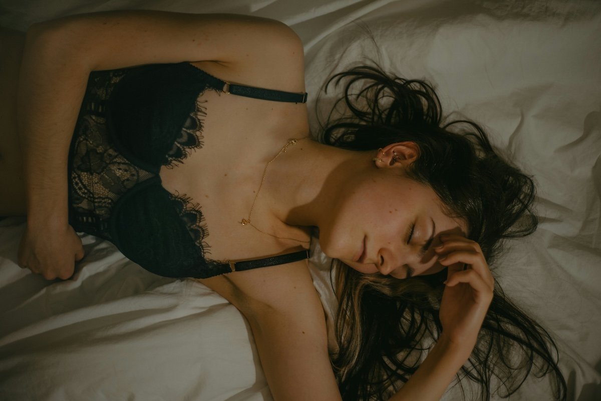 Overhead shot of a woman lying in bed wear black lingerie with her eyes close and hand on her forehead as an example for our boudoir photography guide