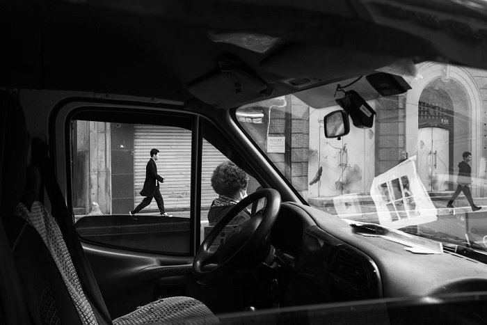 Black and white image shot through an empty car window or man walking on pavement.