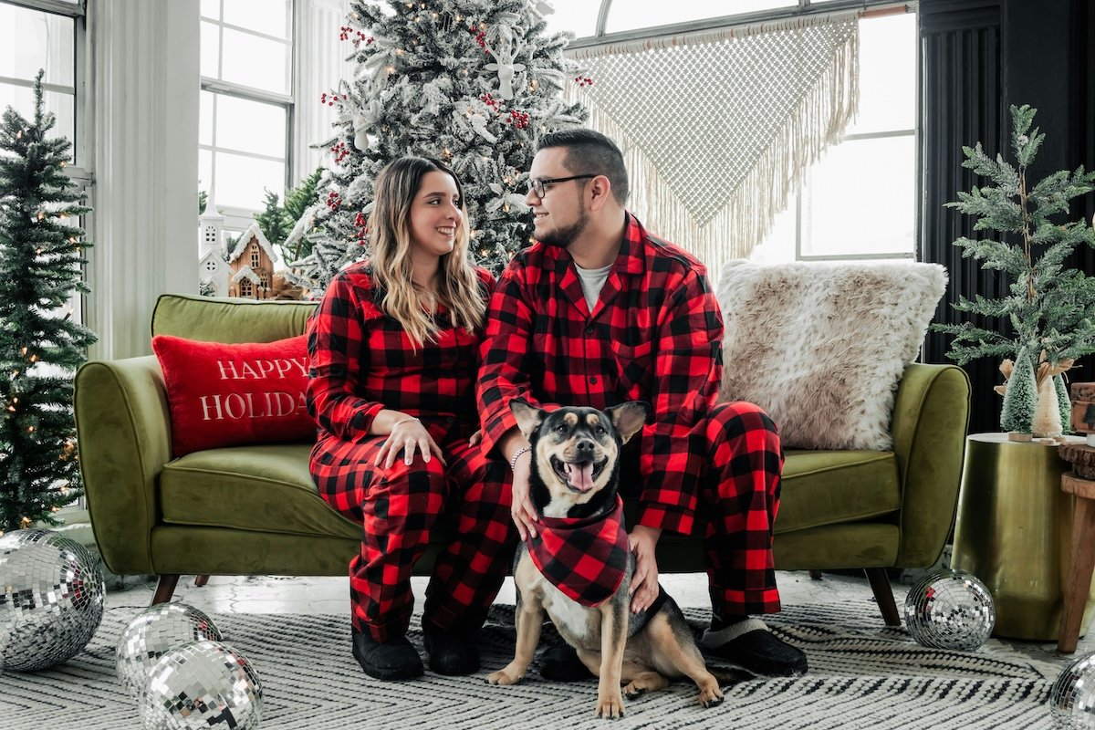A couple posing in onesies on a couch with a Christmas tree in the background and a dog in front as an example of pet photography