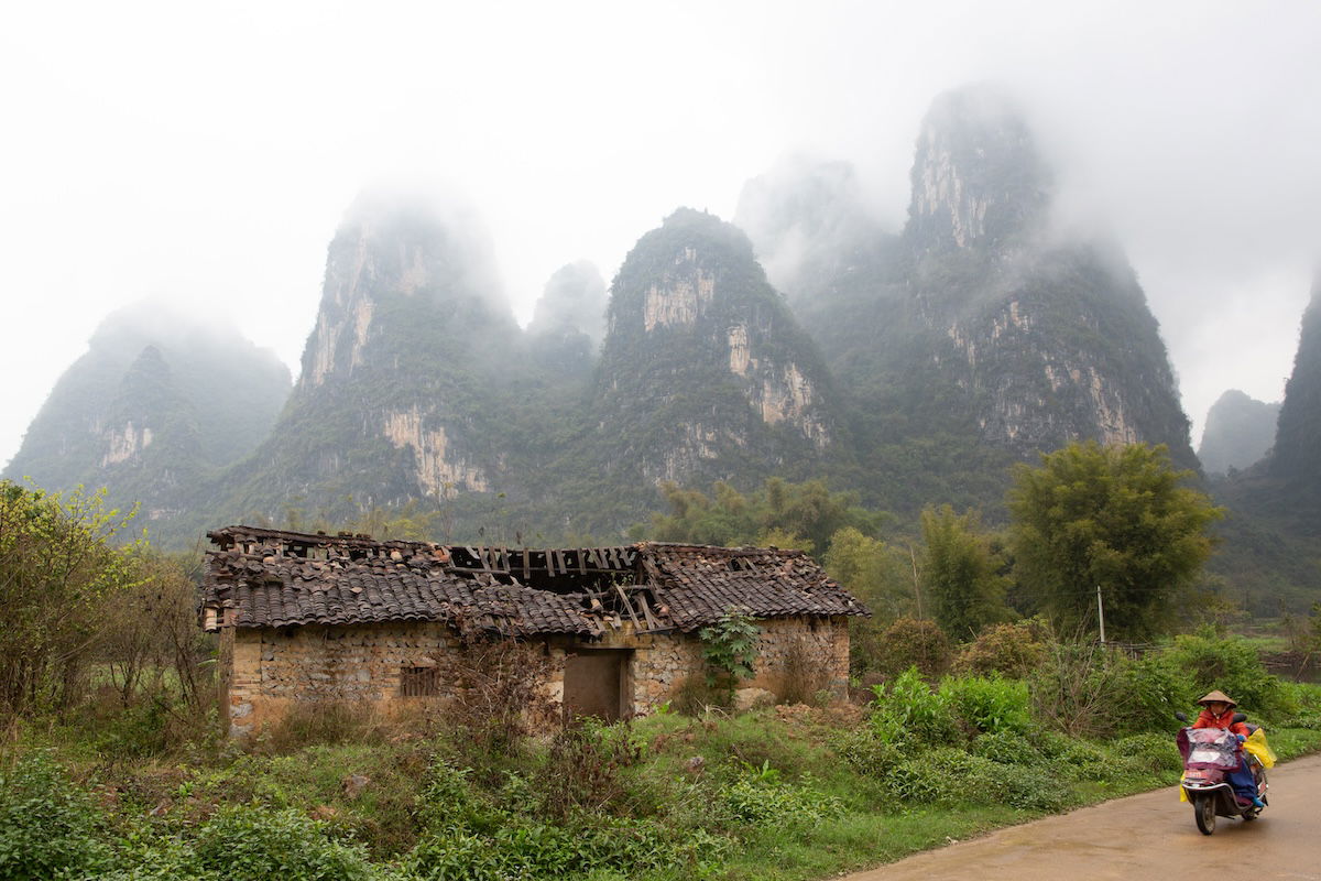 Photograph of mountains in the mist with a abandoned shed in foreground and person on a moped