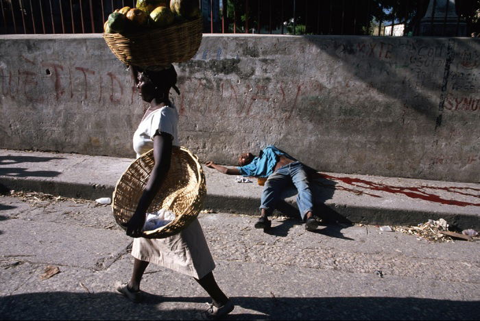 Woman carrying basket of fruit on her head walking past a dead man