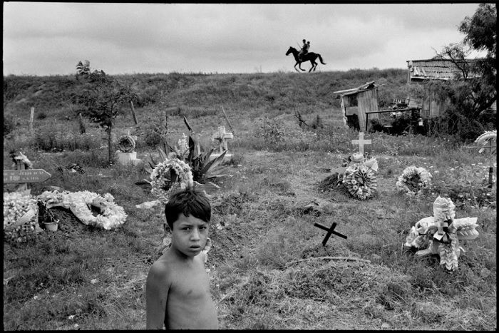 Topless boy in front of poor graveyard with horseman riding in background