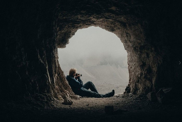 Photographer sitting in the mouth of a cave holding camera to face